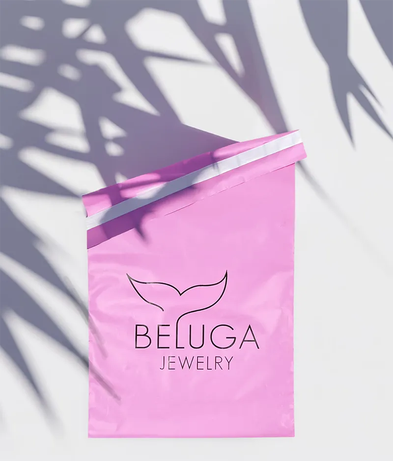 A vibrant pink poly mailer bag is laid flat on a light-colored surface, possibly white or light gray. The bag has a white tear strip visible along its top edge. In the center of the bag, the "BELUGA JEWELRY" logo is printed in black. The logo features a stylized outline of a whale's tail above the word "BELUGA," with "JEWELRY" written in a smaller font below it. The surface around the bag is dappled with strong, clear shadows cast by tropical leaves, suggesting bright sunlight.