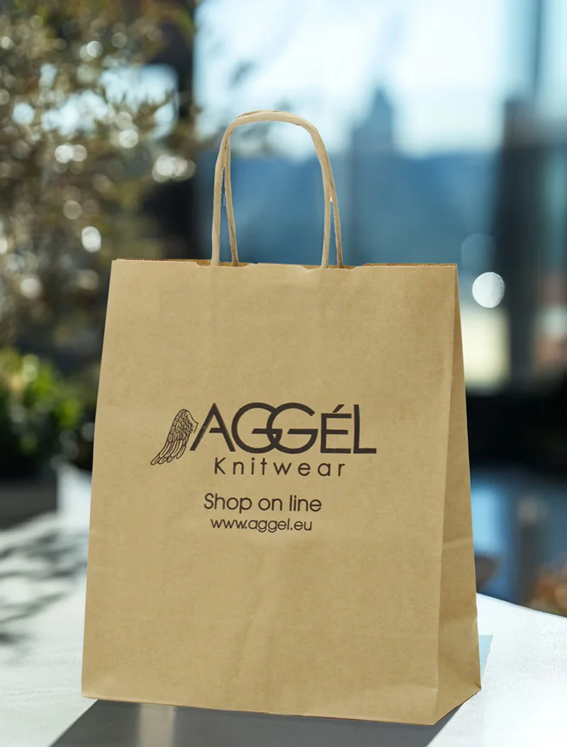 A brown kraft paper shopping bag stands upright on a light-colored surface, likely indoors with a blurred background showing a window and hints of greenery. The bag has brown twisted paper handles. The "AGGÉL KNITWEAR" logo is printed in black on the front, featuring a stylized wing graphic next to the word "AGGÉL." Below the logo, it reads "Shop on line" and "www.aggel.eu" in smaller black text.